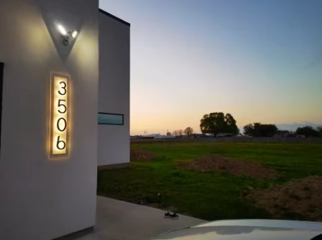 Matte black custom metal LED house numbers illuminated on a modern white exterior wall at dusk