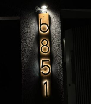 Matte black custom metal LED house numbers mounted on wood panel, illuminated for outdoor use at night.