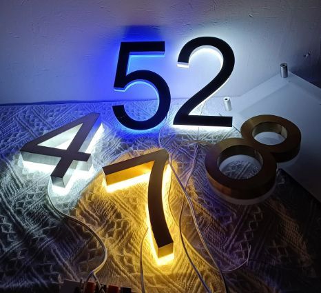 Matte black and brushed metal LED house numbers illuminated with blue, white, and warm yellow backlighting on a table.