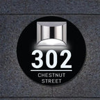 Circular solar-powered acrylic house sign with custom laser-etched address mounted on dark textured wall